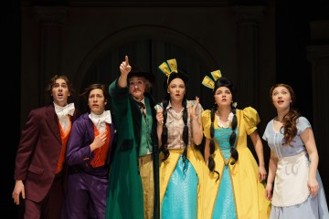 Kuusi värikkäisiin pukuihin pukeutunutta näyttelijää seisoo yhdessä lavalla ja näyttää yllättyneeltä ja huolestuneelta. Yksi osoittaa eteenpäin, kun muut katsovat keskittyneesti samaan suuntaan. He näyttävät olevan keskellä dramaattista kohtausta teatterinäytelmästä.