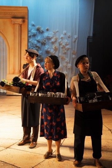 Lavalla seisoo kolme vintage-tyylisesti pukeutunutta ja barettipäistä ihmistä, joilla kullakin on kädessään tarjottimia, joissa on erilaisia esineitä. Taustalla on koristeltu kaari ja seinä, jossa on valkoisia kukka-aksentteja, joita valaisee lämmin näyttämövalaistus.