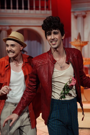 Kaksi värikkäisiin asuihin pukeutunutta miestä hymyilee lavalla. Toisella on punainen takki, ristikaulakoru, kädessään vaaleanpunainen ruusu ja kiharat hiukset. Toisella on olkihattu, punainen paita ja raidalliset housut. Taustalla on suurenmoinen, koristeellinen rakennus.