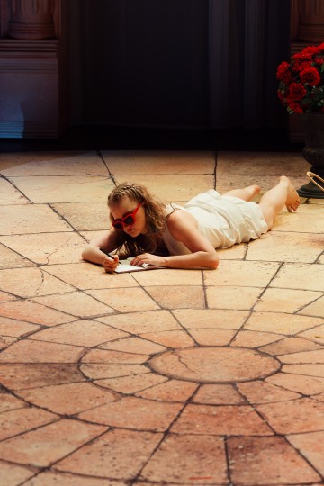 Nainen, jolla on punaiset sydämenmuotoiset aurinkolasit ja valkoinen mekko, makaa pyöreällä kaakelilattialla ja kirjoittaa paperille. Suuri maljakko, jossa on punaisia kukkia, on asetettu lähelle pylväiden ja verhojen muodostamaa taustaa vasten.