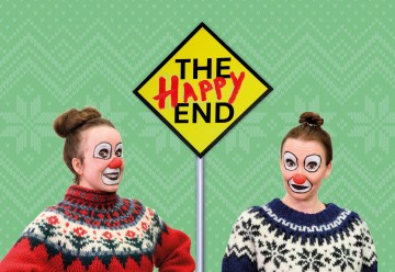 Kaksi värikkäisiin villapaitoihin pukeutunutta klovniksi pukeutunutta naista seisoo hymyillen keltaisen timantinmuotoisen kyltin edessä, jossa lukee 