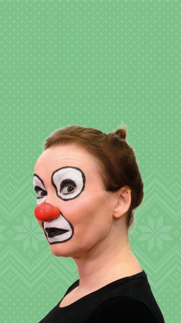 Henkilö, jolla on punainen klovnin nenä ja rohkea mustavalkoinen klovnimeikki, poseeraa profiilissa vihreällä kuviotaustalla, päällään musta toppi ja hiukset taaksepäin sidottuina.