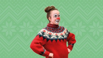 Pelleksi pukeutunut henkilö, jolla on kasvomaali ja punainen nenä, pukeutuu juhlavaan punaisen ja sinisen kuvioiseen puseroon ja seisoo kädet lanteilla vihreällä taustalla, jossa on neulottu lumihiutalekuvio.