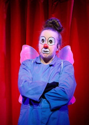 Punaisen verhon edessä seisoo klovniksi pukeutunut henkilö, jolla on punainen nenä, valkoinen kasvomaali ja ilmeikäs meikki, sininen puku ja valaistut keijusiivet, kädet ristissä ja yllättynyt ilme.