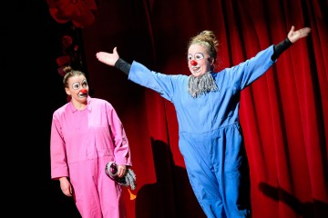 Kaksi pelleä, joilla on punaiset nenät ja kasvomaalit, seisoo punaisen näyttämöverhon edessä. Toinen vaaleanpunaisessa haalarissa pitelee moppia, kun taas toinen sinisessä haalarissa levittää kätensä leveälle ja hymyilee.