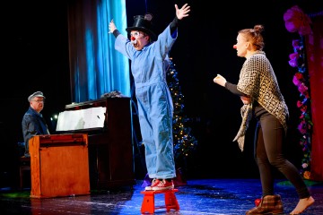 Siniseen haalariin ja silinterihattuun pukeutunut klovni seisoo punaisella jakkaralla kädet ylhäällä ja esiintyy lavalla. Toinen henkilö, jolla on huivi ja punainen nenä, on vuorovaikutuksessa klovnin kanssa. Taustalla soittaa pianisti koristellun joulukuusen vieressä.