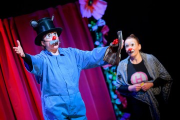 Lavalla esiintyy kaksi esiintyjää värikkäissä puvuissa ja pellemeikissä. Toinen on pukeutunut hiireksi, jolla on sininen puku ja musta silinterihattu, ja hän elehtii vilkkaasti. Toinen katselee yllättyneenä, päällään kuviollinen huivi. Taustalla on punaisia verhoja ja kukkia.