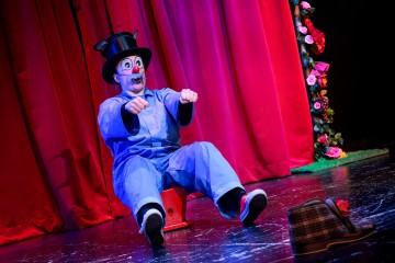 Siniseen haalariin, punaiseen nenään ja silinterihattuun pukeutunut klovni istuu lavalla mimiikkana punaisen verhon edessä. Verhon reunalla on värikkäitä kukkia ja lattialla lähellä on suuri klovnikenkä.