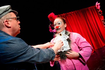 Lakkipukuinen henkilö säätää vaaleanpunaiseen takkiin pukeutuneen, ylimitoitetuilla silmälaseilla ja meikillä varustetun klovniksi pukeutuneen esiintyjän röyhelökaulusta, joka seisoo punaisen, kukilla koristellun verhon edessä.