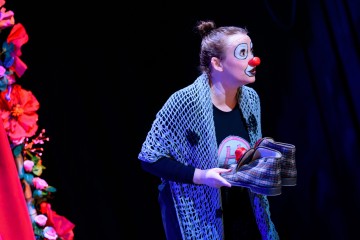 Pelleksi pukeutunut nainen, jolla on punainen nenä ja valkoinen kasvomaali, pitää kädessään paria ruutukenkiä, hänellä on sininen virkattu huivi ja hän seisoo suurten punaisten kukkien vieressä tummaa taustaa vasten.