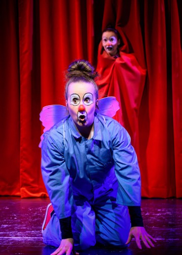 Sinipukuinen esiintyjä, jolla on klovnimeikki ja siivet, polvistuu lavalla suu yllätyksestä auki, kun toinen punapukuinen henkilö, jolla on samanlainen meikki, seisoo hänen takanaan, molemmat punaisen verhon edessä.