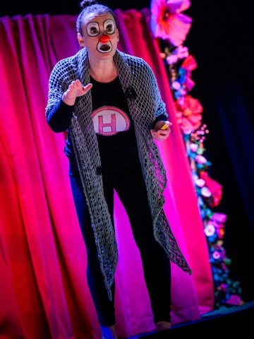 Esiintyjä, jolla on kasvomaali, verkkohuivi ja paita, jossa on suuri H-kirjain, elehtii ilmeikkäästi lavalla, kun hän seisoo vaaleanpunaisen, suurilla, värikkäillä kukilla koristellun verhon edessä.