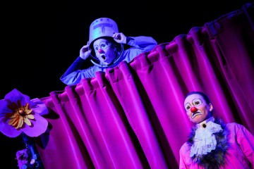 Kaksi klovnia esiintyy lavalla dramaattisessa violetissa valaistuksessa; toinen kurkistaa verhon yli hattu ämpäri päässään, kun taas toinen seisoo edessä surullinen ilme ja röyhelöinen kaulus. Verhoa koristaa suuri kukka.