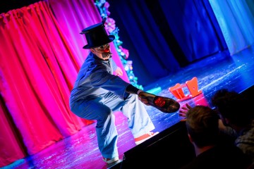 Siniseen pukuun, silinterihattuun, tekoviiksiin ja punaiseen nenään pukeutunut esiintyjä kumartuu yleisöä kohti ja ojentaa kenkää. Lava on kirkkaasti valaistu, ja taustalla on punaisia ja sinisiä verhoja ja rekvisiittaa.