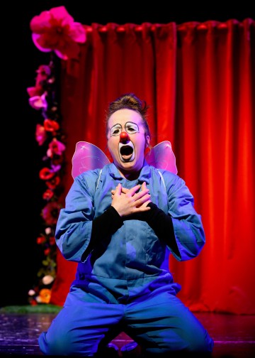 Siniseen haalariin ja keijusiipiin pukeutunut esiintyjä polvistuu lavalla kädet rintakehällä, suu auki. Taustalla on punainen verho ja kukkakoristeita.