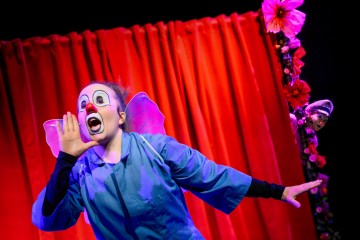 Klovniksi pukeutunut esiintyjä, jolla on kasvomaali, punainen nenä ja vaaleanpunaiset perhossiivet, elehtii dramaattisesti kirkkaanpunaisen, kukkien koristeleman verhon edessä.