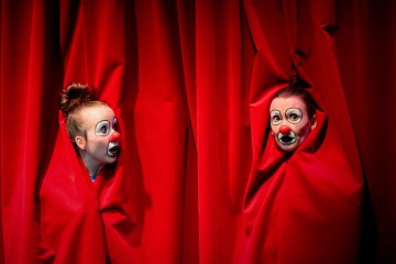 Kaksi valkoisella kasvomaalilla ja punaisella nenällä varustettua klovnia kurkistelee kirkkaanpunaisten näyttämöverhojen läpi, ja heidän kasvonsa ovat kankaan kehystämät ja ilmaisevat liioiteltua yllätystä.