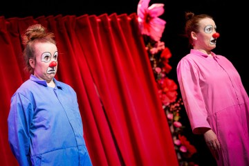 Kaksi klovniksi pukeutunutta esiintyjää, joilla on punaiset nenät ja ilmeikkäät kasvomaalaukset, seisovat punaisten verhojen ja kukkien edessä, toinen sinisessä ja toinen vaaleanpunaisessa asussa, molemmat vakavana.