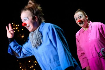 Lavalla seisoo kaksi klovneiksi pukeutunutta ihmistä, joilla on valkoiset kasvomaalit ja punaiset nenät. Toinen siniseen asuun pukeutuneista elehtii kädellään, kun taas toinen vaaleanpunaiseen pukeutunut hymyilee taustalla. Heidän takanaan on pieniä valoja.