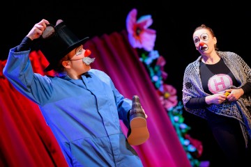 Lavalla esiintyy kaksi klovneiksi pukeutunutta esiintyjää. Toisella on sininen takki, silinterihattu, punainen nenä ja tekoviikset, ja hänellä on kädessään ylisuuret kengät. Toinen, huiviin ja H-paitaan pukeutunut, katselee ilmeikkäästi. Taustalla on vaaleanpunaisia verhoja ja kukkia.