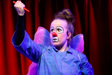 Klovniksi pukeutunut henkilö, jolla on valkoinen kasvomaali, punainen nenä ja perhossiivet, nostaa näyttämöllä dramaattisesti punaista kynää punaisia verhoja vasten.