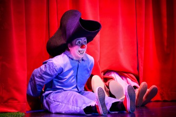 Vaaleansiniseen pukuun ja ylisuureen mustaan hattuun pukeutunut klovni istuu lavalla punaisten verhojen edessä ja hymyilee leveästi. Hänen vieressään verhojen takaa näkyvät vain toisen henkilön jalat ja kengät.