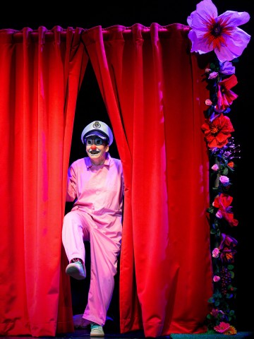 Vaaleanpunaiseen pukuun ja valkoiseen kapteeninhattuun pukeutunut esiintyjä astuu kirkkaanpunaisten, suurilla punaisilla ja violeteilla kukilla koristeltujen näyttämöverhojen läpi ja hymyilee eloisasti näyttämövalaistuksessa.