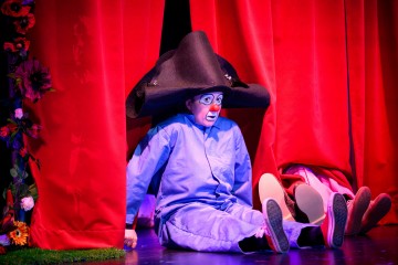 Siniseen pukuun ja ylisuureen mustaan hattuun pukeutunut klovniksi pukeutunut henkilö istuu lavan lattialla ja nojaa kirkkaanpunaisiin verhoihin. Kaksi kenkiin puettua jalkaa työntyy verhojen takaa esiin klovnin vierestä.