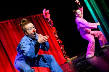 Kaksi esiintyjää kirkkaissa sinisissä ja vaaleanpunaisissa puvuissa, joissa on maalatut klovnin kasvot, esiintyy vilkkaasti lavalla kukkien koristelemien punaisten verhojen edessä. Toinen kyykistyy kädet yhdessä, kun taas toinen seisoo toinen käsi kohotettuna.