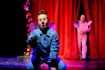 Sinipukuinen esiintyjä, jolla on maalattu klovnimeikki ja keijusiivet, polvistuu dramaattisesti lavalla, kun taas toinen vaaleanpunainen esiintyjä, jolla on kädessään Amerikan lippu, kurkistelee taustalla olevien punaisten verhojen takaa.