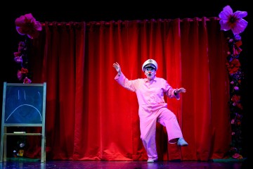 Valkoiseen kapteeninhattuun ja vaaleanpunaiseen asuun pukeutunut esiintyjä poseeraa koomisesti lavalla punaisen verhon edessä, jonka molemmin puolin on kukkia ja liitutaululla surulliset kasvot.