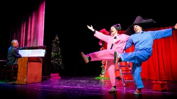 Kaksi klovneiksi pukeutunutta esiintyjää, joilla on vaaleanpunaiset ja siniset puvut ja suuret hatut, potkii jalkojaan lavalla, kun pianisti soittaa vieressä. Taustalla on punainen verho ja koristeltuja joulukuusia.