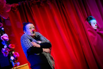 Kaksi esiintyjää, joilla on valkoiset kasvomaalit ja punaiset pellen nenät, seisovat kirkkaanpunaisten verhojen edessä. Toinen, harmaaseen huiviin kääritty, näyttää säikähtäneeltä. Toinen kurkistaa ulos verhon takaa, osittain piilossa.