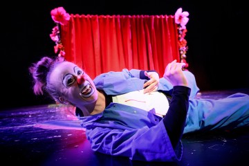 Valkoisella kasvomaalilla, punaisella nenällä ja sinisellä puvulla varustettu klovni makaa naureskellen lavalla. Heidän takanaan on kukkien koristama punainen verho, kaikki kirkkaasti valaistuna tummaa taustaa vasten.