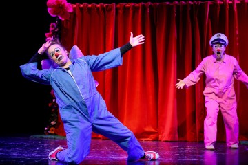 Lavalla esiintyy kaksi esiintyjää värikkäissä puvuissa ja pellemeikissä. Toisella on sininen haalari ja violetit siivet, ja hän nojaa dramaattisesti eteenpäin, kun taas toinen, vaaleanpunaiseen pukeutunut ja kapteeninhattuinen, seisoo kirkkaan punaisen verhon lähellä taustalla.