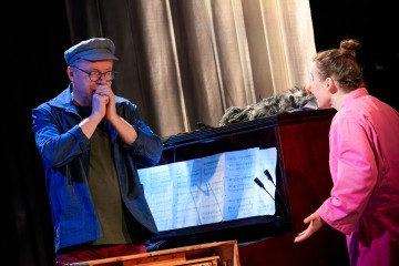 Lakkipäinen mies seisoo nuotteja sisältävän pianon vieressä pitäen käsiään suullaan, kun vaaleanpunaiseen asuun pukeutunut nainen, jolla on punainen pellen nenä, elehtii vilkkaasti häntä kohti.