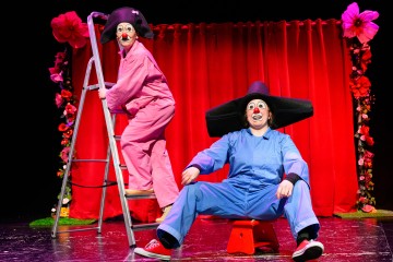 Kaksi punanenäistä klovnia esiintyy lavalla punaisen verhon edessä; toisella on vaaleanpunainen haalari ja hän seisoo tikkailla, kun taas toisella on sininen haalari ja suuri musta hattu, ja hän istuu pienellä punaisella jakkaralla.