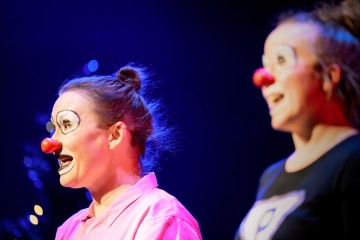 Kaksi pelleiksi pukeutunutta naista, joilla on punaiset nenät ja liioiteltu meikki, esiintyy lavalla kirkkaassa, värikkäässä valaistuksessa. Toisella on vaaleanpunainen paita, toisella musta. Molemmat vaikuttavat eloisilta ja ilmeikkäiltä.