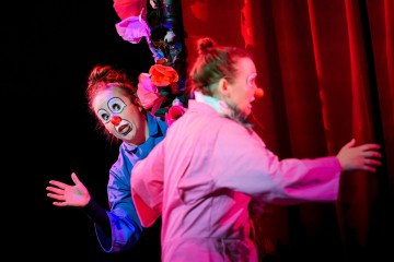 Lavalla esiintyy kaksi pelleä, joilla on punainen nenä ja valkoinen kasvomaali. Toinen kurkistelee yllättyneenä koristellun tolpan takaa, kun taas toinen, vaaleanpunaisessa takissa, siirtyy poispäin kädet ylhäällä. Punaiset verhot ja kukat ovat näkyvissä.