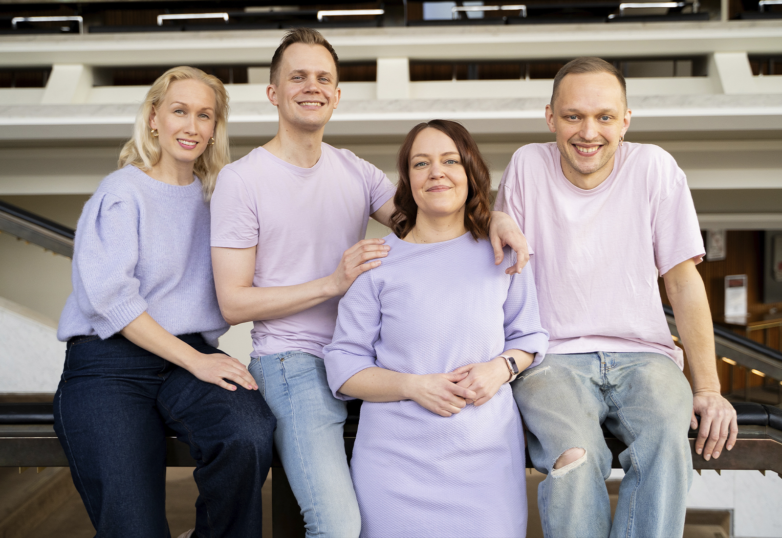 Neljä aikuista poseeraa yhdessä sisätiloissa, ja kaikilla on vaalean violetit tai laventelinväriset topit ja farkut. He hymyilevät ja seisovat lähellä toisiaan valoisassa, modernissa ympäristössä.