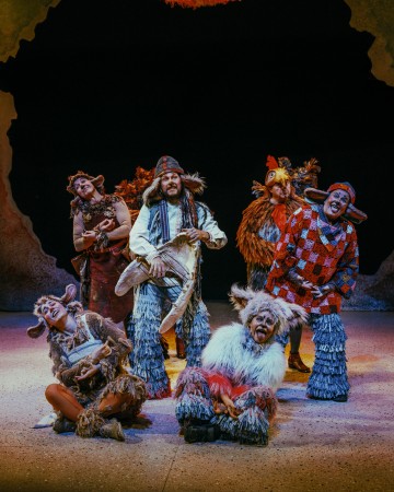 Kuusi näyttelijää pukeutuu näyttämöllä värikkäisiin, omituisiin eläinpukuihin, kuten turkiksiin, sarviin ja höyheniin, ja näyttelijät poseeraavat eloisasti ja leikkisästi luolamaista näyttämötaustaa vasten.