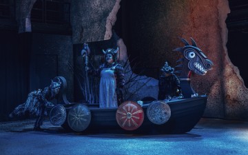 Lavalla seisoo kolme viikinkiaikaisiin asuihin pukeutunutta esiintyjää lohikäärmeenpäisessä puuveneessä, jonka kyljessä on kilvet. Kohtaus on valaistu sinisin sävyin, ja kallioinen, dramaattinen tausta luo teatterillisen ja myyttisen tunnelman.