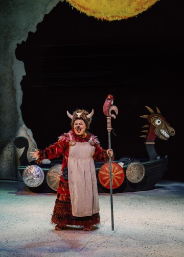 Sarvipäiseen kypärään, punaiseen pukuun ja beigeen esiliinaan pukeutunut esiintyjä laulaa intohimoisesti lavalla sauva kädessään. Heidän takanaan on viikinkilaivan lavaste, jossa on värikkäitä kilpiä ja tyylitelty hevosen pää.