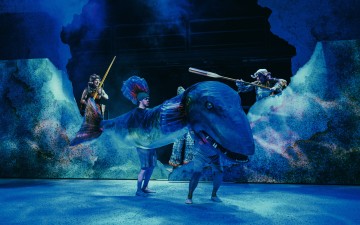 Näyttelijät pitelevät meloja ja käyttävät suurta sinistä valaan nukkea, jota ympäröi sininen valaistus ja vettä muistuttavat lavasteet, jotka luovat dramaattisen valtamerikohtauksen.