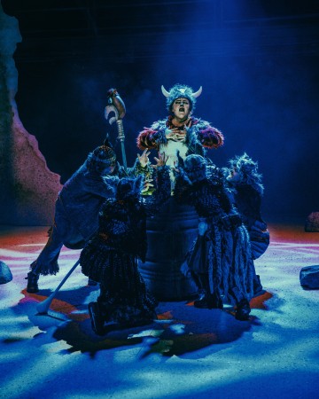 Sarvipäiseen viikinkikypärään pukeutunut esiintyjä seisoo näyttämön keskellä, dramaattisesti valaistuna, ja häntä ympäröi neljä ihmistä, jotka ovat pukeutuneet tummiin pukuihin ja naamioihin. Sininen ja violetti näyttämövalaistus luo mystisen tunnelman.