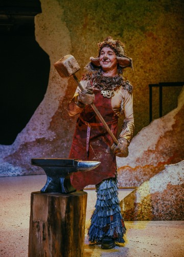 Henkilö, jolla on omituinen puku, johon kuuluu karvainen hattu ja esiliina, hymyilee pitäessään suurta vasaraa alasin yläpuolella puupölkyn päällä, seisoo lavalla, jossa on värikäs, kuvioitu tausta.