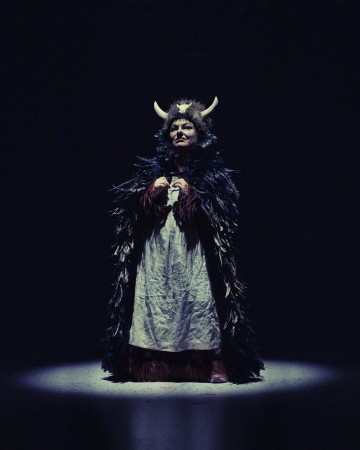 Henkilö seisoo valonheittimen alla yllään dramaattinen puku, jossa on mustia höyheniä, pitkä valkoinen mekko ja sarvipäinen päähine tummalla taustalla.