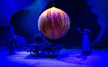 Kolme värikkäisiin pukuihin pukeutunutta esiintyjää ympäröi suurta, hehkuvaa, moniväristä palloa, joka on ripustettu puukärryn yläpuolelle sinisellä valaistulla näyttämöllä, jonka taustalla on abstrakteja maisemia.