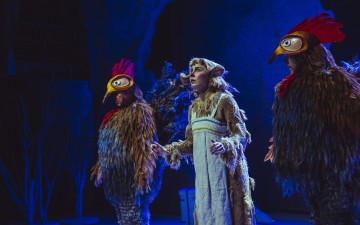 Lavalla kolme näyttelijää; kahdella on värikkäät, höyhenpeitteiset lintupuvut, joissa on suuret punaiset kammat, kun taas keskimmäisellä näyttelijällä on eläimelliset korvat ja turkisasu, kaikki dramaattisen sinisen näyttämövalaistuksen alla.