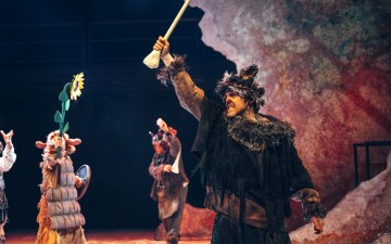 Eläinmäiseen turkispukuun pukeutunut mies nostaa dramaattisesti lavalla tekoluuta, ja muut näyttelijät esiintyvät omituisissa eläinasuissa taustalla kallioisen lavastuksen edessä.